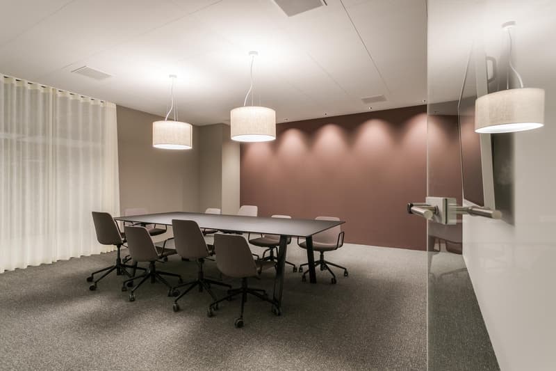Large conference table with 8 chairs, pendant lighting fixtures, gray carpeted floor, gray and pink accent walls