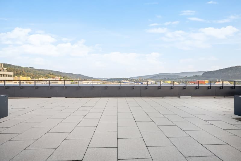 The image shows a large outdoor plaza or terrace with a paved floor made of gray concrete tiles. The plaza overlooks a cityscape with buildings and mountains in the background, under a partly cloudy blue sky.