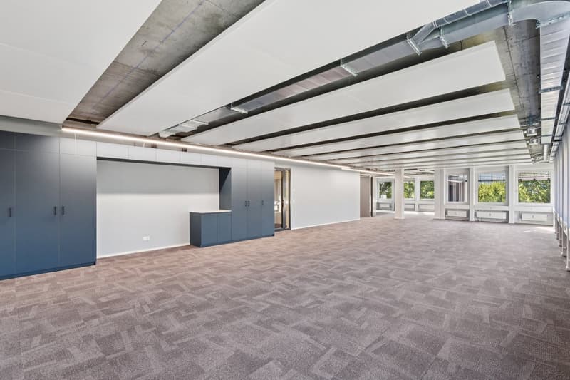 Large open office space with exposed ceiling beams, gray carpeted flooring, and built-in storage cabinets. The space has large windows providing natural light and a modern, industrial-style design.