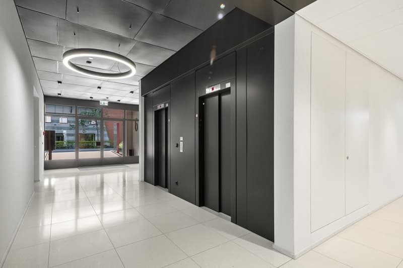 The image shows a modern and minimalist lobby area of a building. The space features a high ceiling with a circular lighting fixture, black elevator doors, and a glass entryway that provides a view of the exterior. The floor is covered with white tiles, c