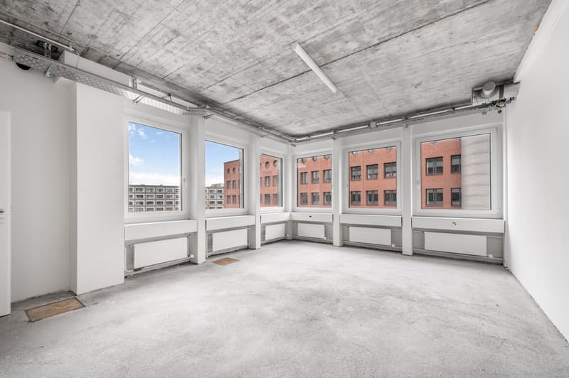 Large open-plan office space with concrete ceiling, white walls, and large windows overlooking surrounding buildings. The room has a bare concrete floor and appears to be in a commercial or industrial building undergoing renovation or conversion.
