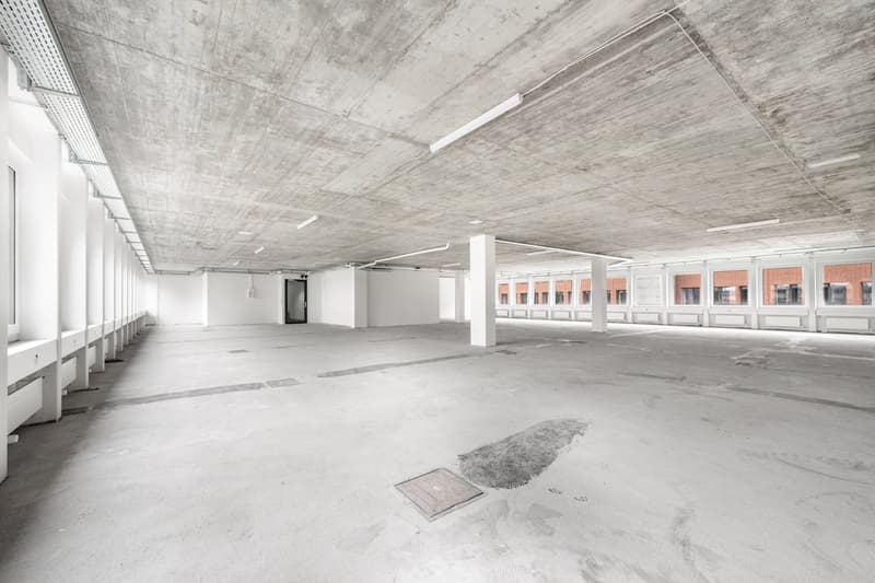 Large open commercial space with concrete ceiling and floor, white walls, and rows of windows along the sides. The space appears to be an unfinished or renovated office or warehouse area.