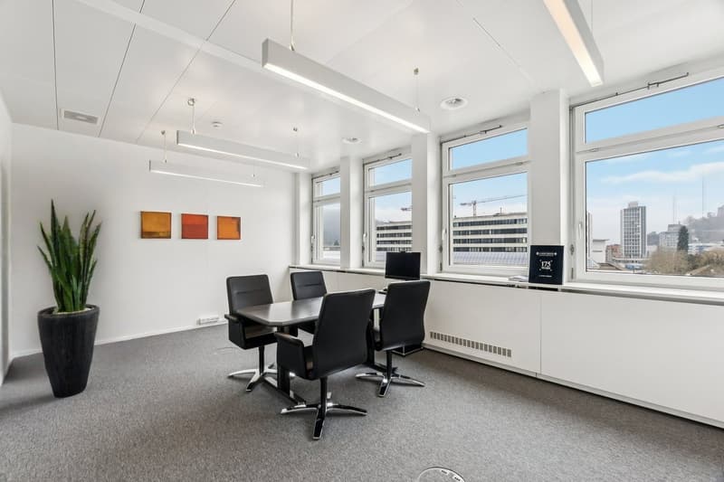 A minimalist office room with white walls, ceilings, and gray carpet, featuring a round dining table with chairs, large windows with a view, and a black plant in a vase.