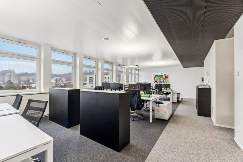 An office space with several workstations, desks, and large windows providing a view of the city.