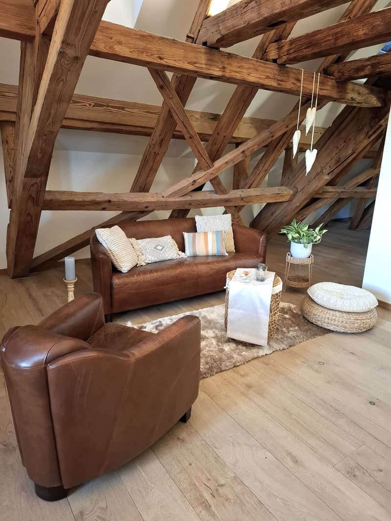 An attic living room features wooden beams and exposed rafters with a wooden floor, a brown leather couch with decorative pillows, a side table with a plant, a round pillow, and a small round table with a white cover and some objects.