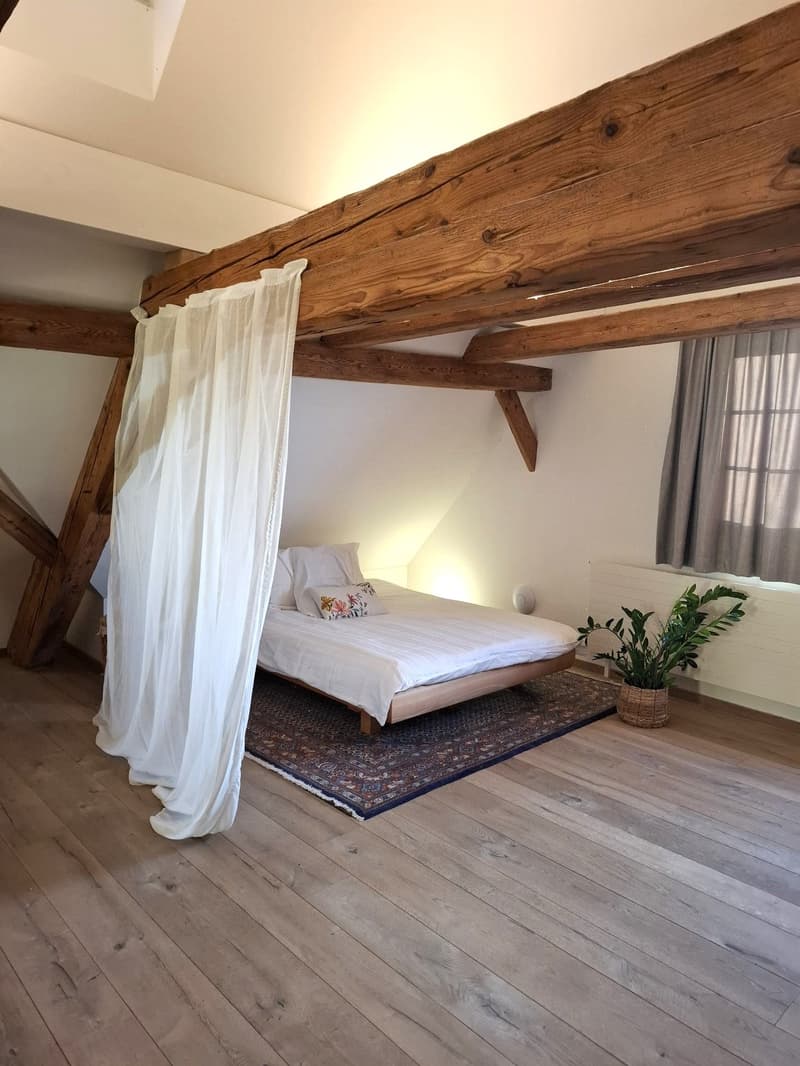 wooden floor, exposed wooden beams, bed with white sheets, pillows, grey rug, white curtains