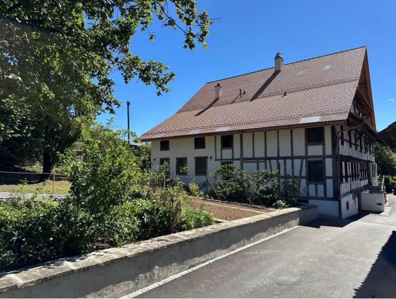 White and brown house, red tiled roof, chimneys, several windows, concrete driveway, large bushes