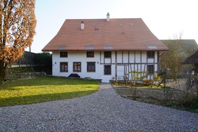 2-storey house with a chimney, red roof, gravel driveway, white walls, multiple windows, solar panels on roof, fenced yard, trees and plants