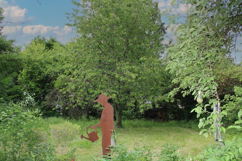 Lush green garden with a large tree, a metal sculpture of a person holding a watering can, and other greenery surrounding the scene.
