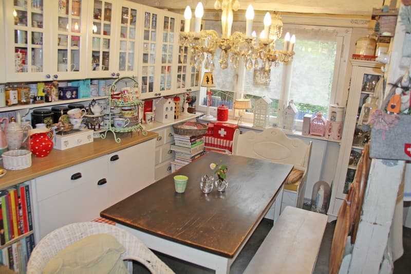 Cozy and eclectic kitchen with glass-front cabinets, a large chandelier, a wooden dining table, and various decorative items and appliances. The kitchen appears to have a homey and personalized feel.