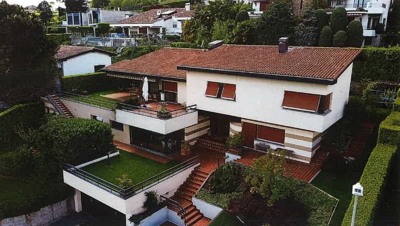 Two-story house with a raised ground floor, balconies, and a parking area. The house has a white exterior with red tile roofing and large windows. The property is surrounded by lush greenery and landscaping.
