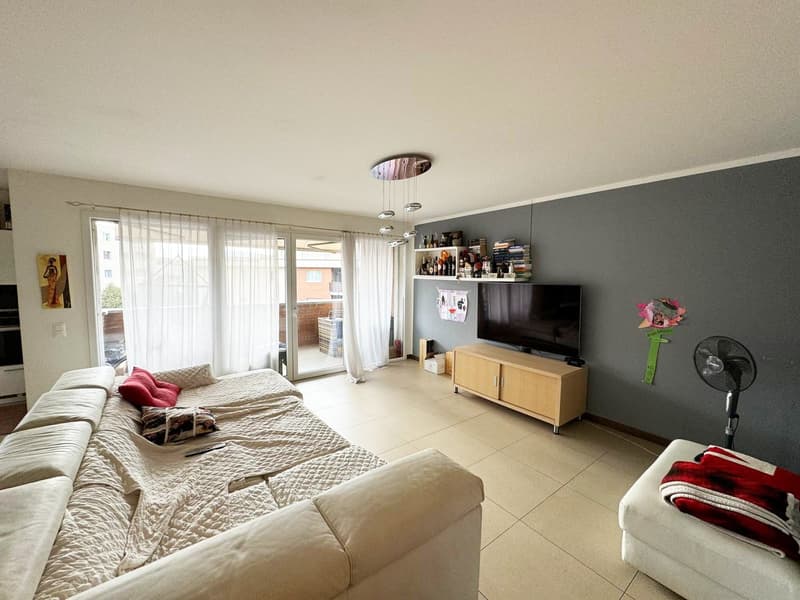 Spacious living room with large windows, beige sofa, TV stand, and shelves with books and decor items. The room has a balcony or terrace visible through the sliding glass doors.