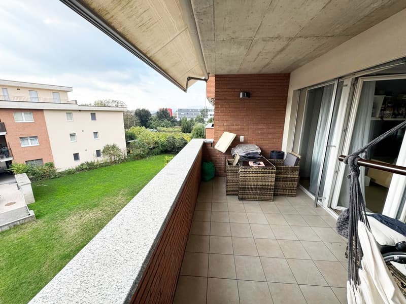 Spacious balcony with concrete ceiling, brick walls, and tile flooring. Wicker furniture and storage boxes on the balcony. View of a grassy area and other apartment buildings outside.