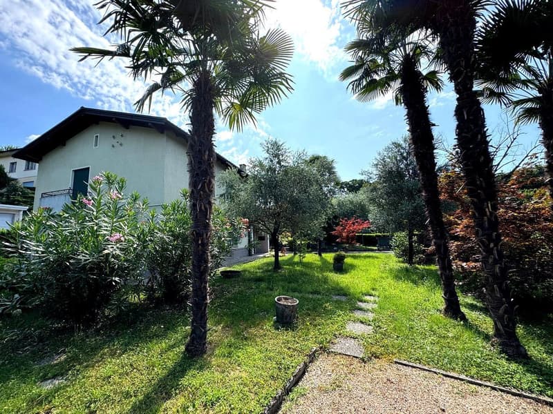 Two-story house surrounded by lush tropical vegetation, including palm trees and other greenery. The house has a patio or balcony area visible. The property appears to have a well-maintained garden with a grassy lawn and a paved walkway.