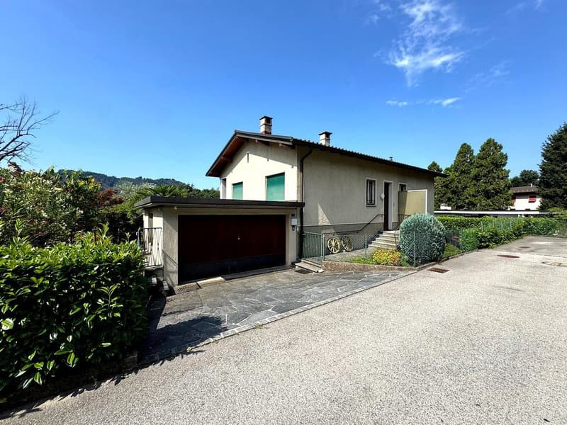 2-story house with a garage, surrounded by lush greenery and a paved driveway. The house has a gray exterior with a tiled roof and chimneys.