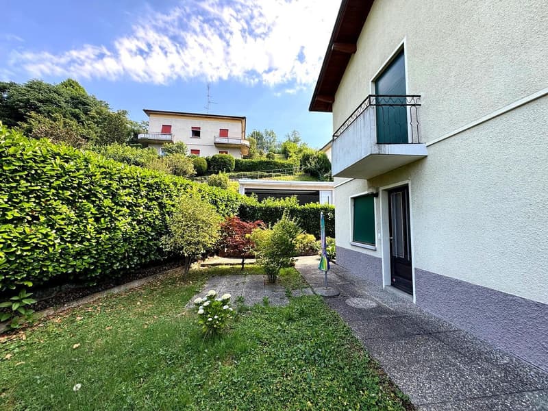 Two-story residential building with a balcony, surrounded by lush greenery and landscaping. The building has a white exterior with red accents and large windows. There is a paved walkway leading to the entrance and a well-maintained garden area with shrub