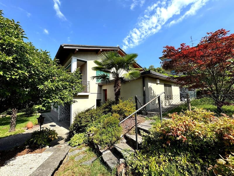 2-story house with a balcony, raised ground floor, garage, and parking space. The house has a tropical garden with palm trees and colorful foliage.