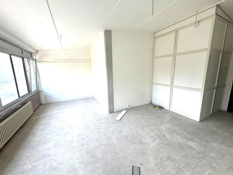 An empty room with white walls, gray floor, large windows, radiators, and some construction materials.