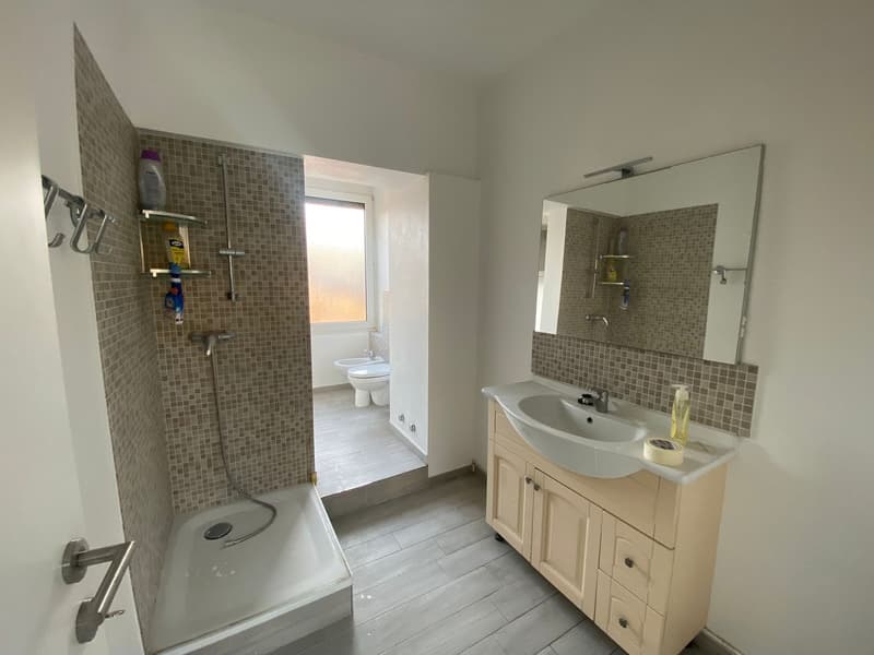 White tile floor, shower area with glass door, sink with wooden cabinet, mirror, toilet area, window, tiled walls
