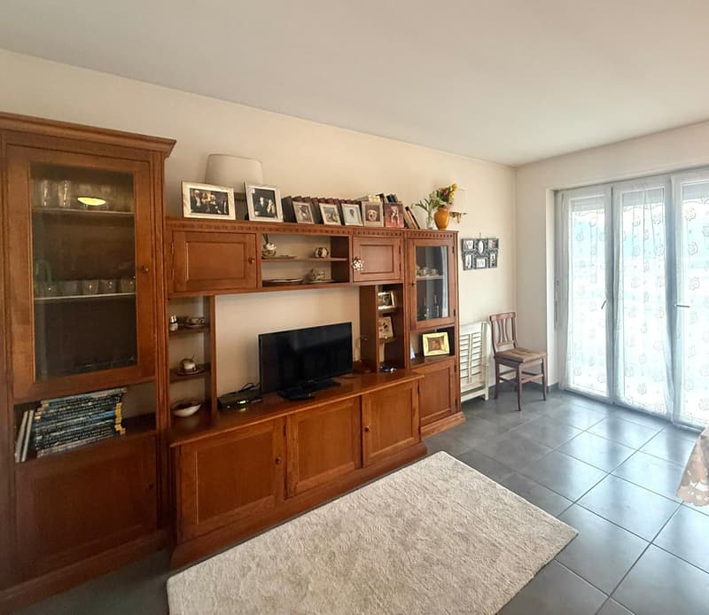 Living room with wooden cabinet, TV, couch, chairs, carpet, table, sliding doors leading to balcony, white curtains