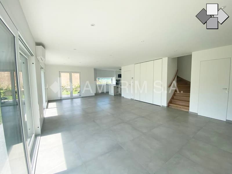 Spacious open-plan living area with large windows, white walls, and grey tile flooring. The room features built-in storage cabinets and a staircase leading to an upper level. There is a sliding glass door that provides access to a balcony or terrace area.
