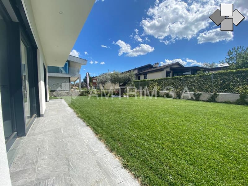 Two-story house with a large yard, paved walkway, and green lawn. The house has a balcony or terrace visible, and there appears to be a parking space or driveway on the property.