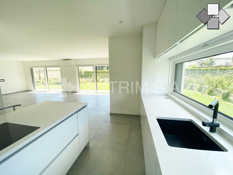 Spacious open-plan kitchen with modern white cabinets, countertops, and large windows overlooking the garden. The kitchen features a dishwasher and a sink with a faucet.