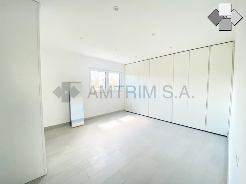 Spacious bedroom with white walls, large built-in wardrobes, and a bright, open feel. The room appears to have a balcony or terrace accessible through a window.