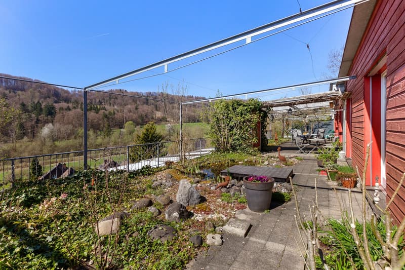 Outdoor area with a wooden deck, metal pergola, plants, and a small pond. The garden is surrounded by a metal fence and has a view of the hills and trees.