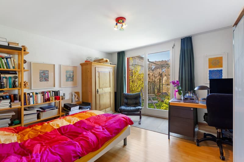 Bedroom with bed, wooden cabinet, desk, chair, bookshelves, and a glass door leading to a balcony with a view of the outside.