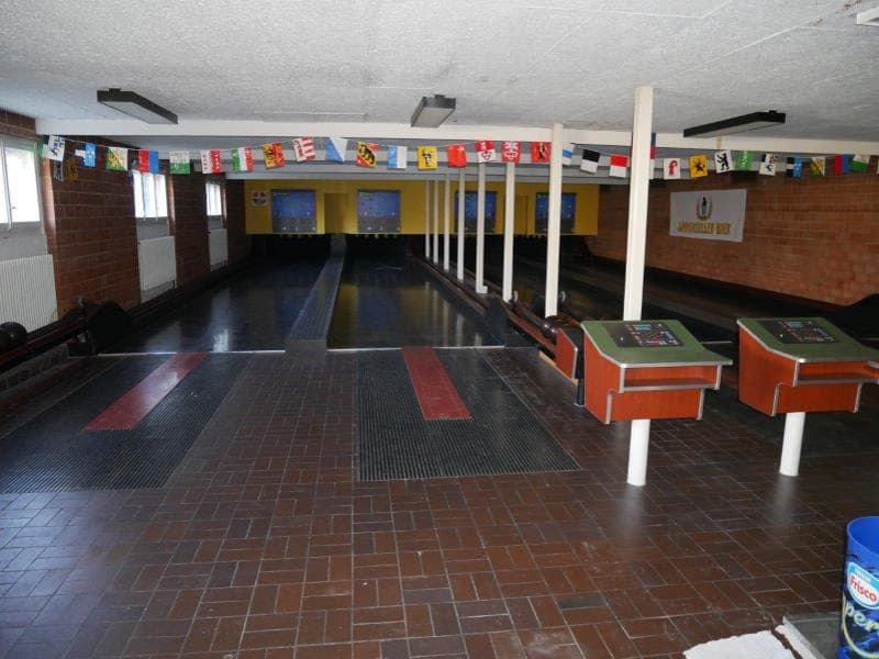 A large indoor bowling alley, featuring multiple bowling lanes, flags on a wall, counters for scores, and radiators, with a sign on the wall