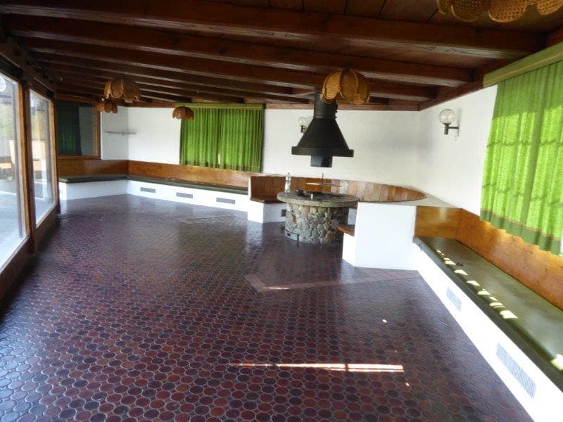 Stone fireplace in the center of the room, large windows, green curtains, wooden benches along the walls, brown tile flooring.