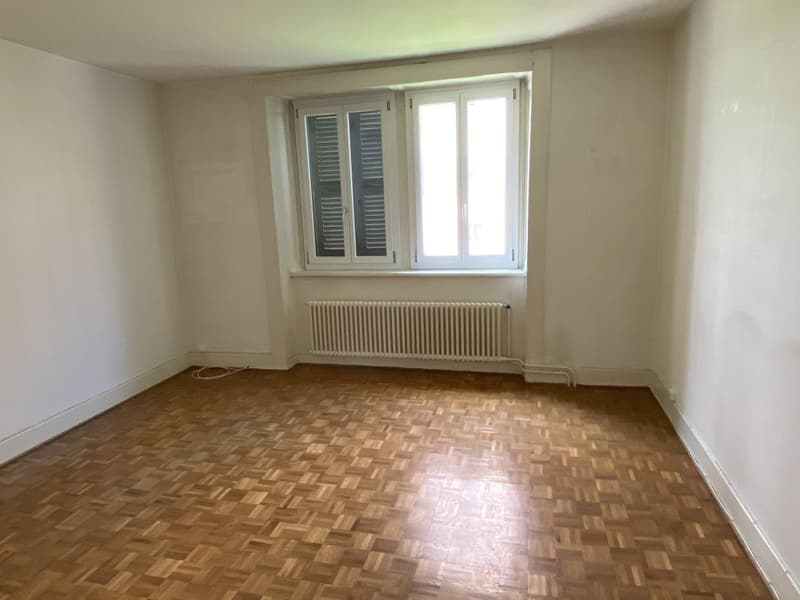 Spacious bedroom with hardwood parquet flooring, white walls, and a large window with shutters. The room appears to be empty, ready for furnishing.