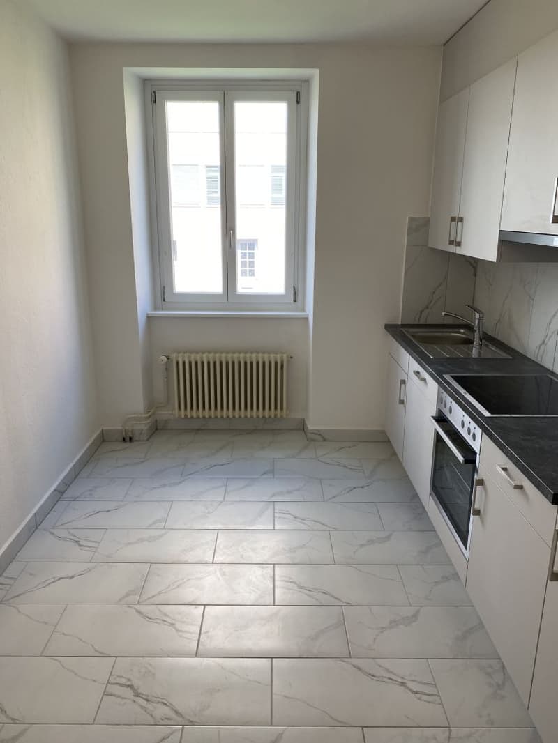 Empty kitchen, white tiles, white cabinets, black countertops, white sink, oven, window with white frame, radiator
