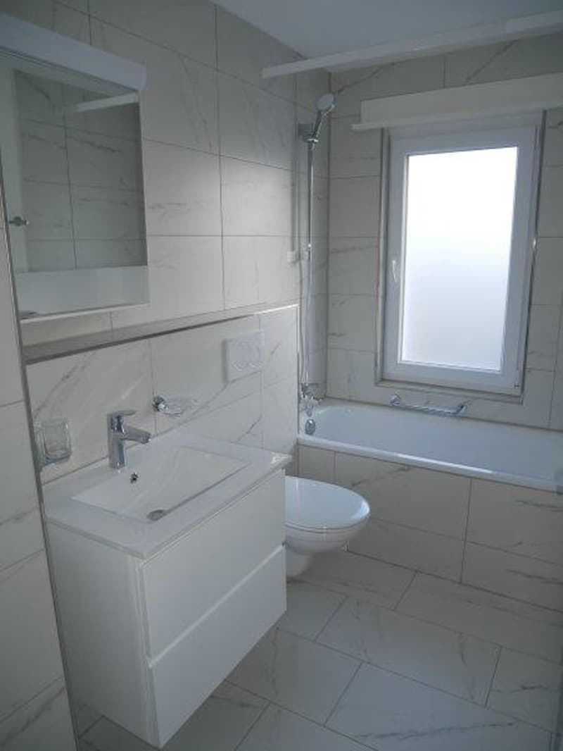 A clean and minimalist bathroom with white tiles, a bathtub, a toilet, a sink, a mirror, and a window. Modern fixtures.