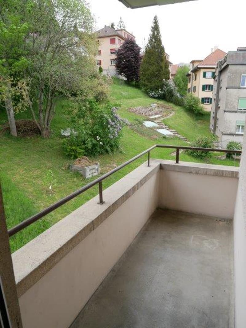 The image shows a balcony or terrace overlooking a grassy area with trees and buildings in the background. The balcony has a concrete floor and a metal railing.