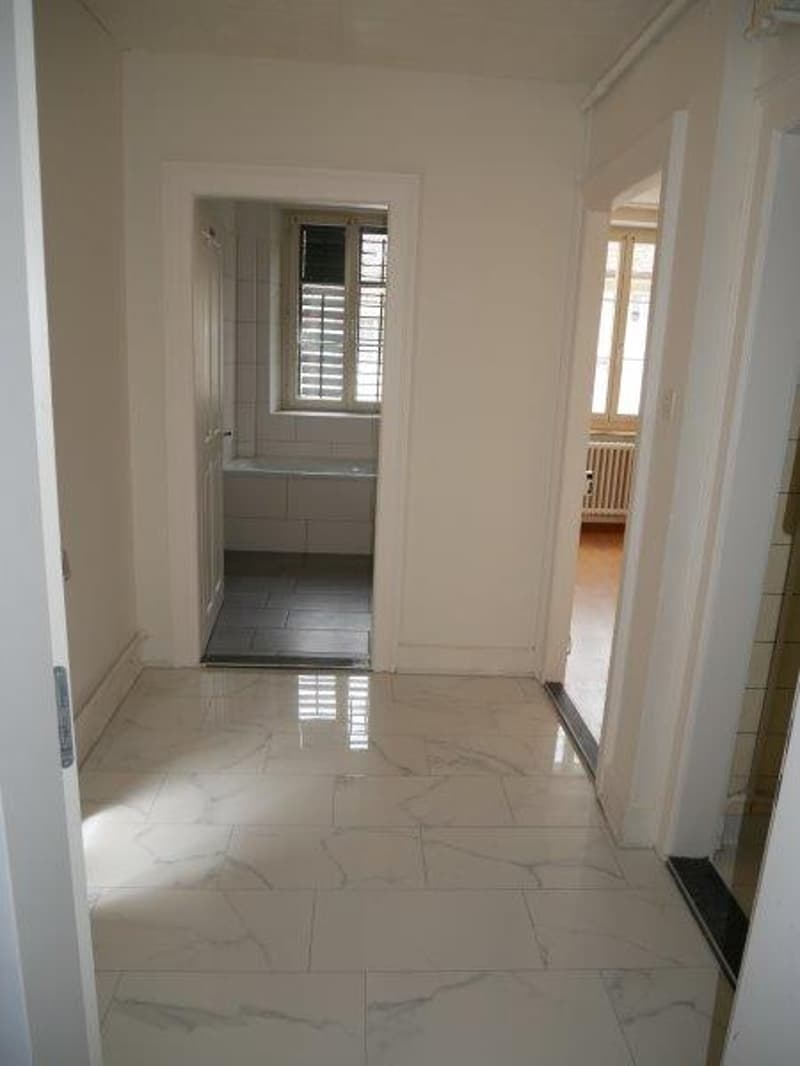 The image shows a hallway with a tiled floor made of large, white marble-like tiles. The walls are painted in a neutral, light color. There is a doorway leading to another room visible at the end of the hallway.