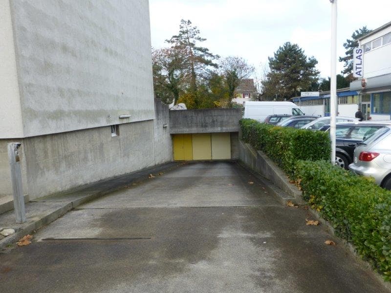 An empty parking lot leads to a yellow garage entrance. There are several cars parked in front of a building. The parking lot is concrete with some dried leaves and has bushes on the side. There is a pole with the word Atlas on it.
