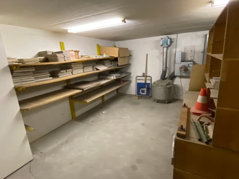 An empty storage room with a wooden shelf and boxes, metal objects, a gray machine, a white wall, a blue sign, a broom, and a traffic cone