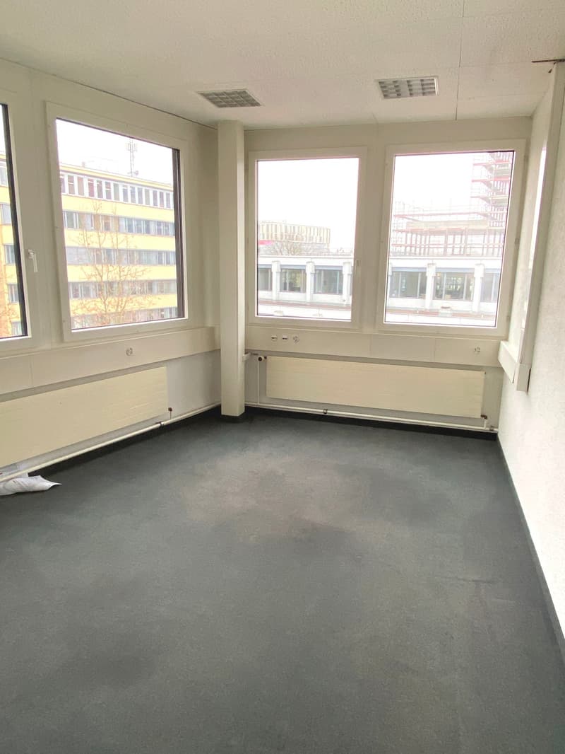 An empty room with three windows, dark gray carpet flooring, three white radiators, and view of the outside.