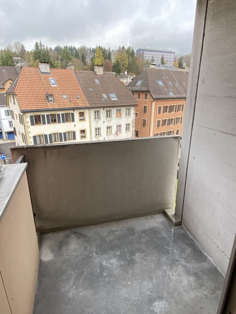 The image shows a balcony or terrace overlooking a cityscape with traditional European-style buildings with red tile roofs and dormer windows. The balcony has a concrete floor and is surrounded by a concrete wall.