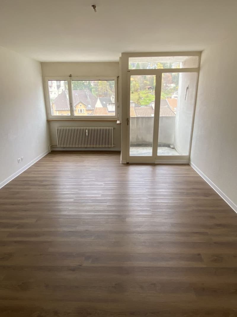 Spacious living room with hardwood flooring, large windows providing natural light, and a sliding glass door leading to a balcony or terrace with a view of the surrounding neighborhood.