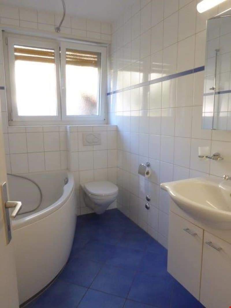 Bathroom with bathtub, toilet, sink, window, blue tile flooring, white walls and cabinets