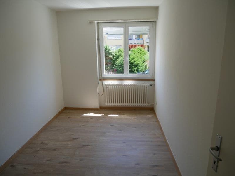 Empty room with white walls, wooden floor, window, radiator, and a door.