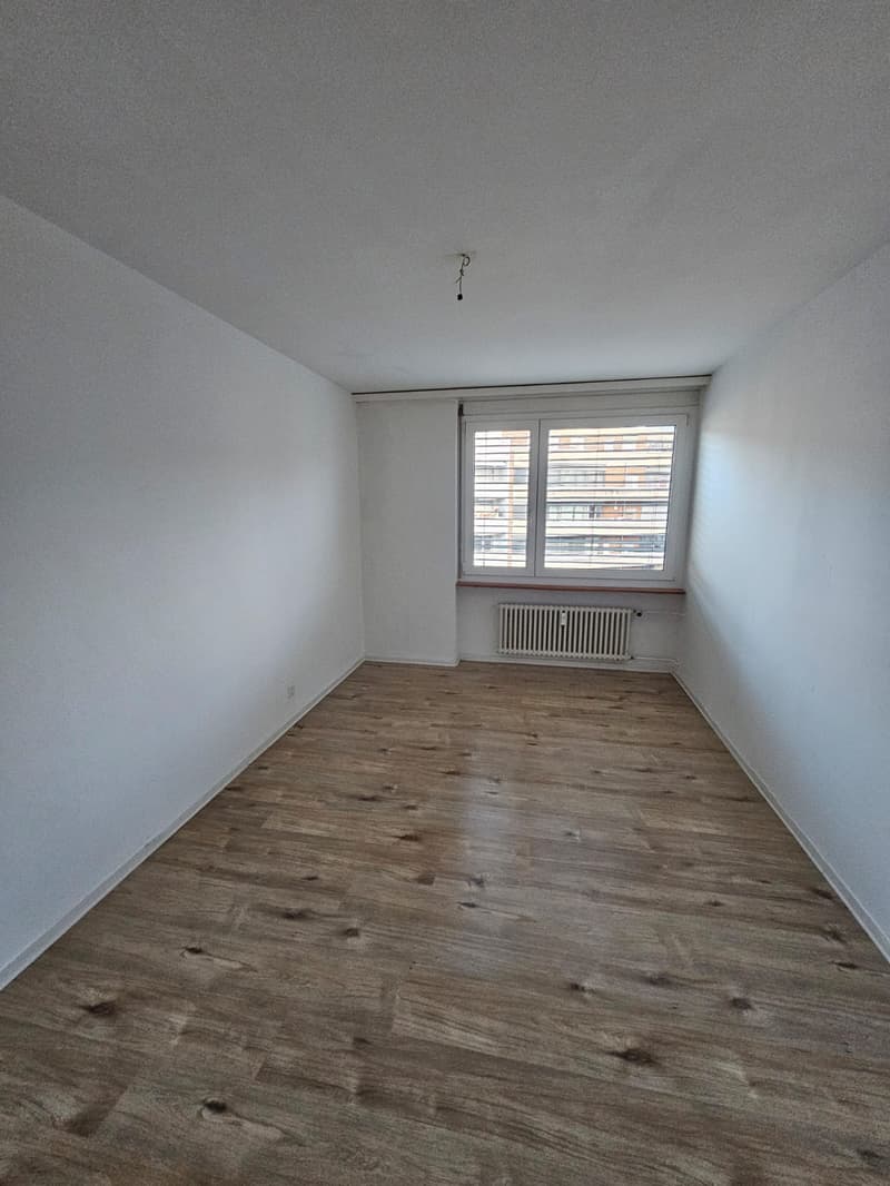 an empty room with white walls, wooden floor, and a window with blinds