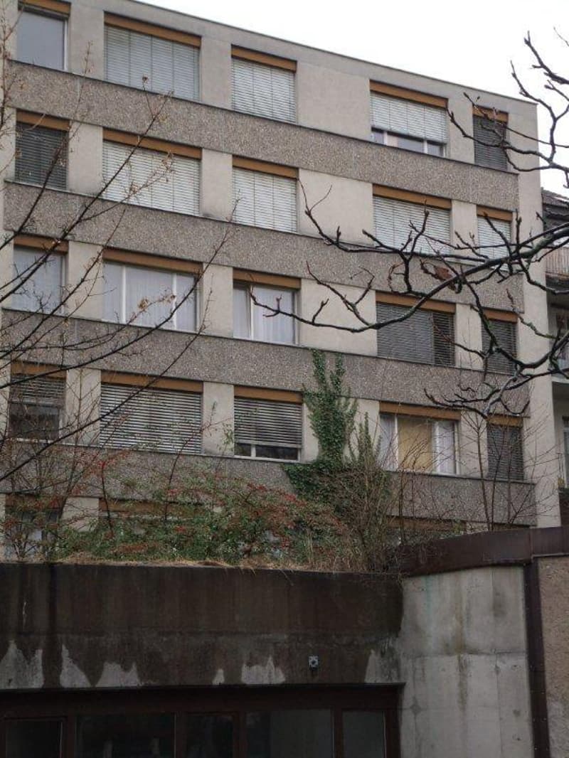 A multi-story building with concrete exterior, multiple windows, some with shutters, a balcony on the top floor, and a green rooftop with plants and bushes.