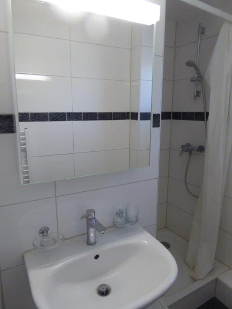A small, modern bathroom with white tiles, a sink, mirror, and a shower. There is also a soap dish, two glasses, and a shower curtain.