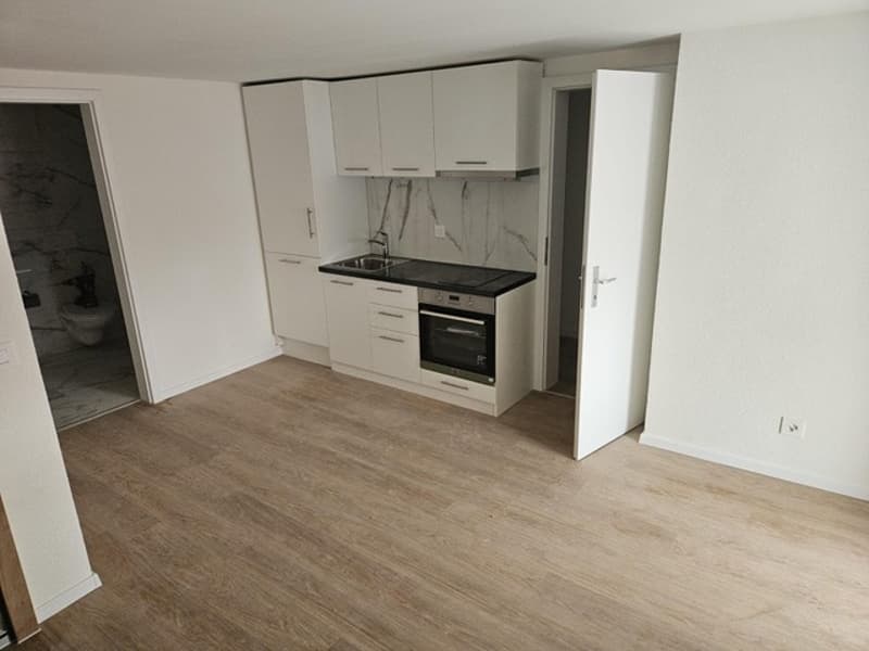 Empty kitchen with white cabinets, wooden floor, black countertop, sink, oven, and a door leading to the bathroom.
