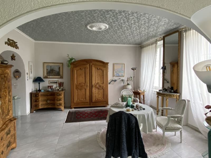 Dining room with wooden furniture, white walls, tiled floor, large window with curtains, table with tablecloth, chairs, cabinet, mirror, and various decorative items