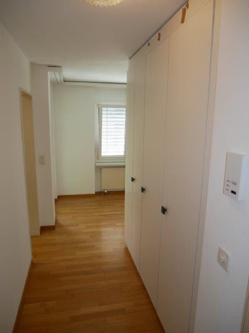 Empty hallway with wooden floor, white walls, built-in wardrobes, window with blinds, power outlets, and a radiator.